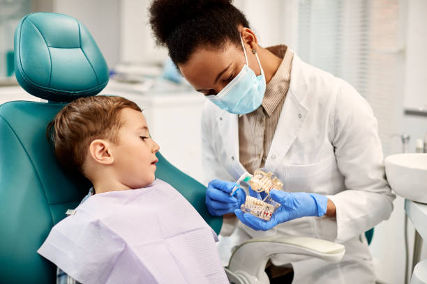 A dentist wearing a facemask holds a toothbrush and dental model to show a child how to brush their teeth.