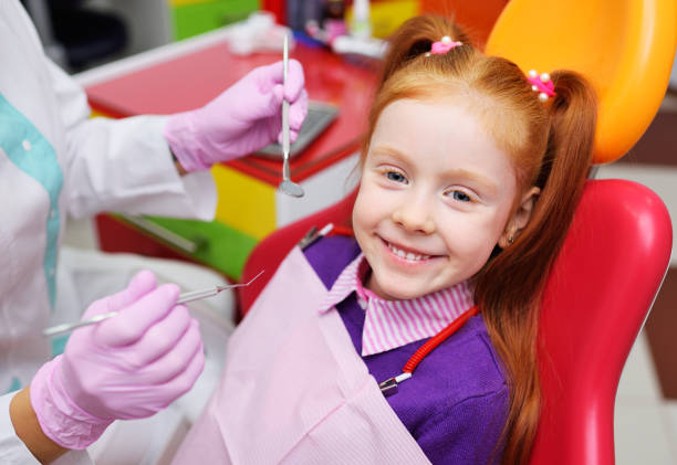 A young child with red hair sits in a red chair and smiles as a dentist prepares to inspect her mouth with dental equipment.