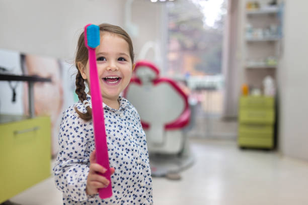 A girl holds an oversized pink tooth brush at her side as she smiles at the camera.