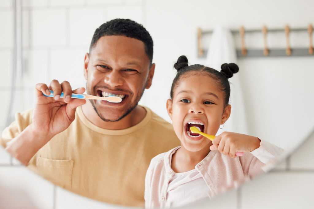 A father and daughter brushing teeth together in the mirror.