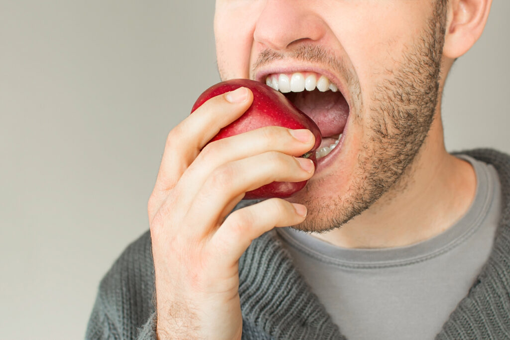 Closeup of a young man with a beard eating an apple on a grey plain background