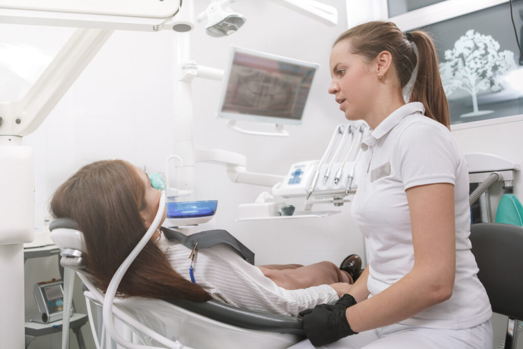 A young woman with a nose mask on listens to a dental hygienist while she gets sedated.